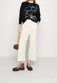 Kobieta w czarnym swetrze z dużym białym wzorem kwiatu, w jasnych szerokich spodniach, brązowych butach za kostkę, trzymająca małą brązową torbę z beżowym paskiem.