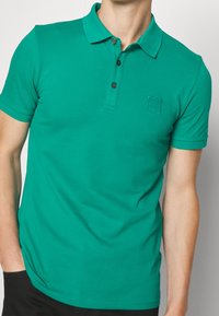 Polo vert avec un plastron à boutons, des manches courtes et un petit écusson logo. Fabriqué en tissu lisse avec une coupe ajustée.