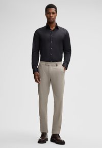 Uomo in piedi che indossa una camicia nera a maniche lunghe, pantaloni beige su misura e stivali in pelle marrone scuro su uno sfondo chiaro e semplice.
