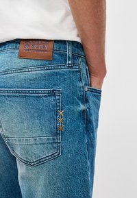 Ανοιχτού μπλε τζιν παντελόνι με δερμάτινη ετικέτα επωνυμίας, πίσω τσέπη με πορτοκαλί ραφή και σχέδιο κανονικής εφαρμογής.