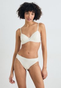Csipkés szegélyű, világos krémszínű bralette állítható pántokkal, amelyhez illő, csipke díszítésű alsónemű tartozik, mindkettő puha anyagból készült.