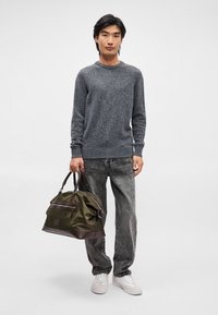 Pulover texturat gri, blugi gri închis și adidași albi. Ținând o geantă verde cu accente maro și un fermoar, care are un buzunar frontal.