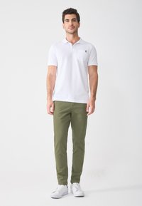 Weißes Polohemd mit kleiner Tasche, olivgrüne Hose und weiße Sneakers. Glatte Textur und figurbetontes Design mit klassischem Schnitt.