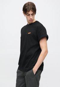 Μαύρο βαμβακερό T-shirt με γυρισμένα μανίκια, με πορτοκαλί λογότυπο στο στήθος. Απλός, άνετος σχεδιασμός.