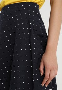 Marinblå kjol med vita prickar, med en utsvängd design och plisserade detaljer. Slät textur och en åtsittande midja.