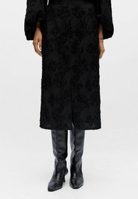 Sort blomstrede teksturerede midi-frakke båret med sorte spidse læderstøvler til knæet, hænder afslappet langs siderne mod en hvid baggrund.