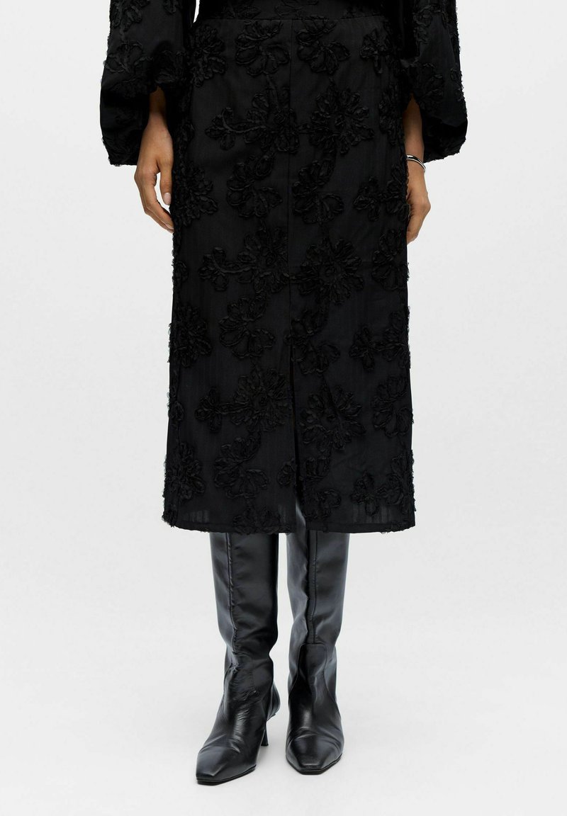 Sort blomstrede teksturerede midi-frakke båret med sorte spidse læderstøvler til knæet, hænder afslappet langs siderne mod en hvid baggrund.