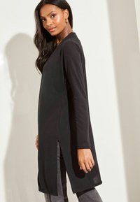 Tunique noire à manches longues avec fentes latérales, en tissu doux. Design simple avec décolleté en V, portée sur un jean gris, sur un fond clair.