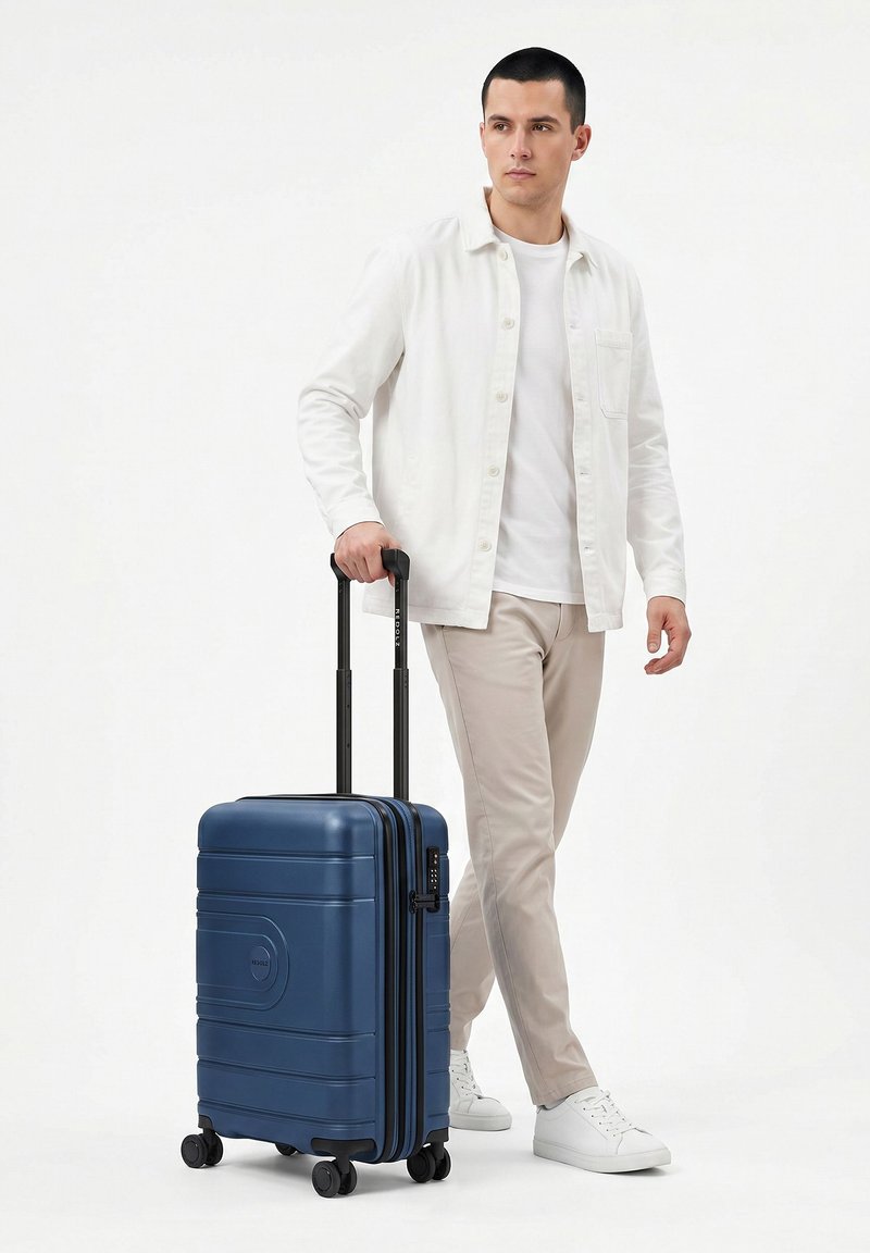 Homme en chemise blanche et pantalon beige marchant en tirant une valise rigide bleue à quatre roues avec une poignée télescopique.