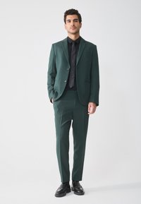 Dunkelgrüner Anzug mit schlanker Passform, einreihigem Blazer und passender Hose. Kombiniert mit einem schwarzen Hemd und einer Krawatte sowie Lederschuhen.