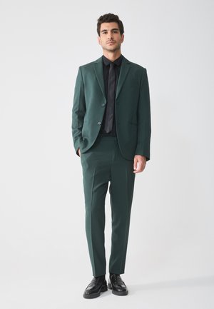 Dunkelgrüner Anzug mit schlanker Passform, einreihigem Blazer und passender Hose. Kombiniert mit einem schwarzen Hemd und einer Krawatte sowie Lederschuhen.