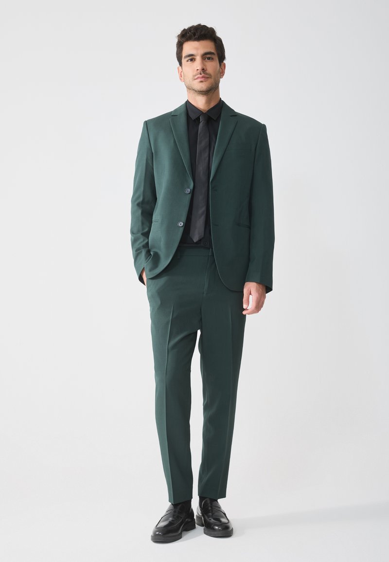 Dunkelgrüner Anzug mit schlanker Passform, einreihigem Blazer und passender Hose. Kombiniert mit einem schwarzen Hemd und einer Krawatte sowie Lederschuhen.