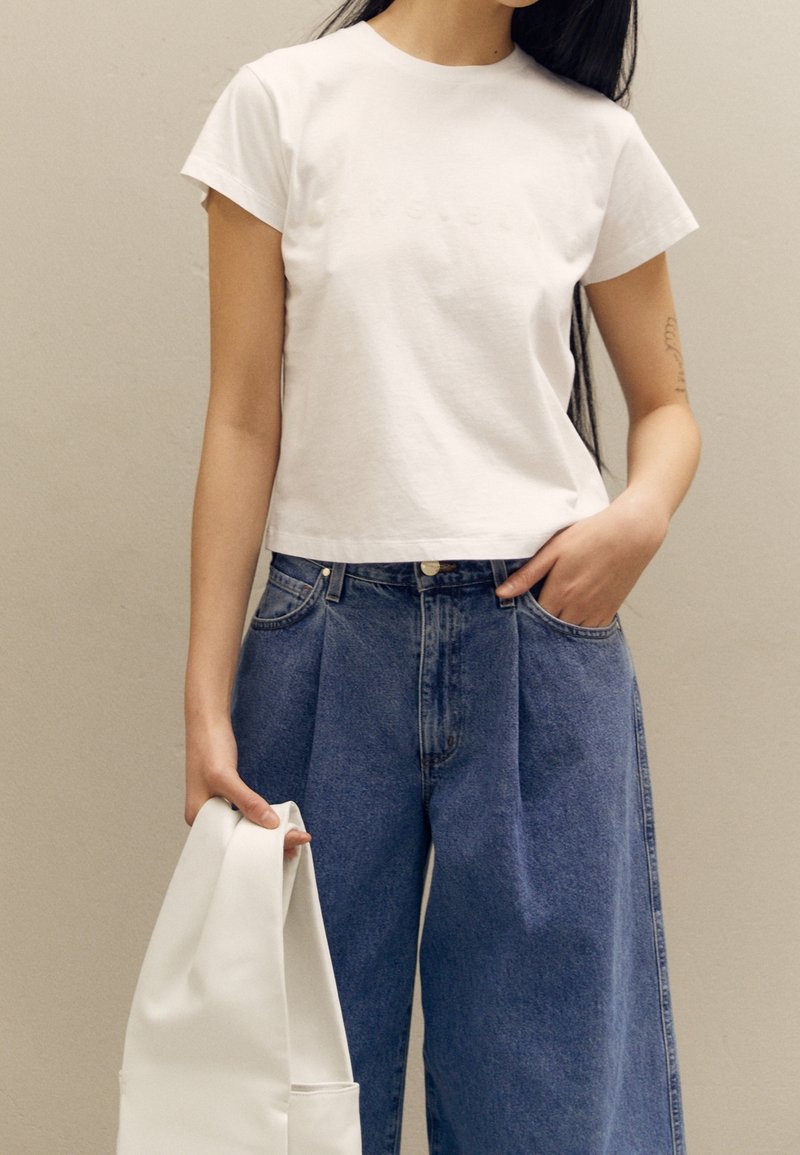 Vit kortärmad t-shirt med präglad text, parat med högmidjade ljusblå denimbyxor. Modellen håller en slät vit väska.