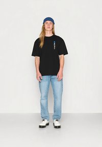 Melna oversize T-krekls ar zilu akcentu un tekstu, kombinēts ar gaiši ziliem džinsiem un baltām kurpēm ar melnām zolēm, valkāts kopā ar zilu cepuri.