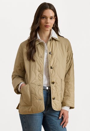 Beige quilted jacket with a button front, two pockets, and a collar. Worn over a white shirt, featuring a relaxed fit and soft fabric texture.