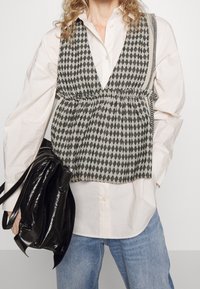 Chaleco a rayas en blanco y negro con cintura elástica, llevado sobre una camisa de botones clara. Bolso negro brillante en la mano. Jeans de mezclilla azul.