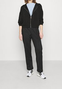 Svart zip-up huva jacka med en slät textur, kombinerad med svarta byxor. Modellen har på sig vita och svarta mönstrade sneakers. Ljusblå skjorta under.