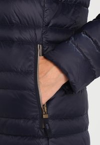 Marinefarbener Puffer-Jacke mit horizontalem Steppdesign, seitlicher Reißverschlusstasche und einem metallischen Reißverschluss. Glattes, leichtes Material mit einem eleganten Aussehen.
