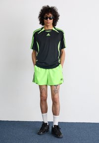 Sort træningstrøje med grønne detaljer, neongrønne shorts, sorte sneakers og solbriller. Har tatoveringer på benene.
