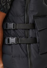 Czarna pikowana kamizelka z centralnym zamkiem błyskawicznym, regulowanymi paskami w talii oraz boczną kieszenią z klapką. Miękka, gładka tekstura.