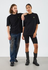 Storede sorte t-shirts med et gult logo, kombineret med denim jeans og sorte cykelshorts. Kvinden bærer høje sorte støvler med detaljer.