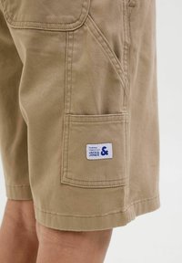 Shorts kaki en tissu résistant, avec des poches latérales et arrière, et une étiquette cousue avec un texte bleu sur la poche arrière.