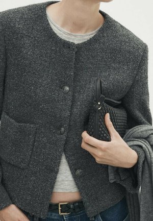 Personne portant une veste texturée gris foncé boutonnée, tenant une pochette noire tissée et un vêtement gris plié, avec un jean et une ceinture visibles.