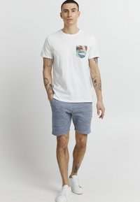 T-shirt en coton blanc avec un design de poche multicolore, associé à un short texturé gris-bleu et à des baskets blanches.