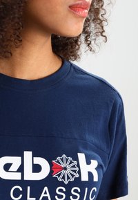 Námořnická modrá bavlněná trička s kulatým výstřihem a krátkým rukávem, na kterém je bílé logo "Reebok" a grafika se zářením.