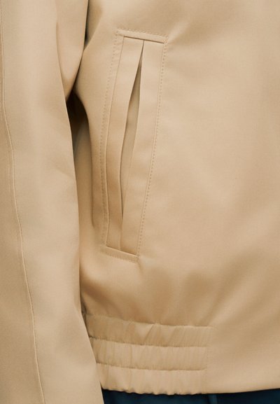 Gros plan sur une poche de veste beige avec une fente verticale et une taille élastiquée en dessous.
