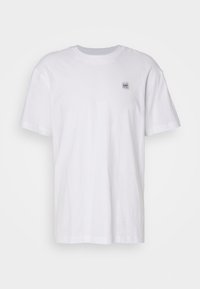 Bílý bavlněný T-shirt s krátkým rukávem, kulatým výstřihem a malou modro-bílou nášivkou na levé hrudi. Normální střih, hladká textura.