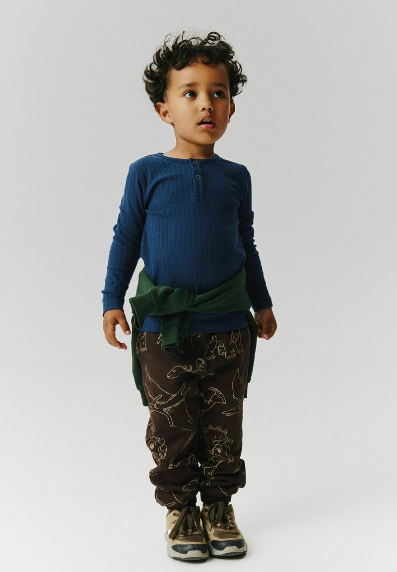 Jeune enfant aux cheveux bouclés portant un t-shirt bleu à manches longues, un pantalon à motif dinosaure, un pull vert noué autour de la taille et des baskets beiges.