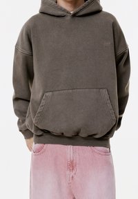 Sweat à capuche marron avec une poche kangourou et des poignets côtelés. Porté avec un jean en denim rose clair, présentant une coupe décontractée et oversized.