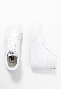 Vita högklackade sneakers i slätt läder med runda tår och vadderad krage. Har en karakteristisk sidovinge och synliga sömmar.