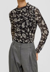 Langärmliges Oberteil mit einem schwarz-grauen Blumenmuster und einem hohen Kragen. Kombiniert mit hochgeschnittenen grauen Hosen, die Falten zeigen.