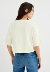 Suéter corto color crema con mangas cortas, de suave textura y sutiles detalles acanalados, combinado con jeans de mezclilla azul.