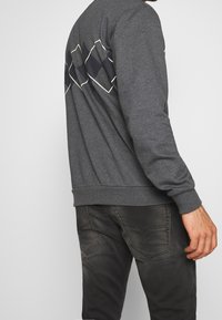 Grå sweatshirt med ett geometriskt diamantmönster i svart och vitt på ryggen, tillsammans med mörka jeans. Mjuk tyg, avslappnad passform.