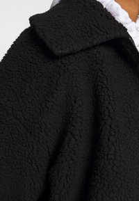 Close-up van een zwarte gestructureerde fleece jas met grote kraag, gedragen over een witte gerimpelde top.