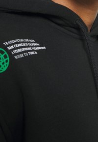 Sudadera negra de tejido suave con un logo de globo verde y detalles en texto blanco. Capucha con cordón, vista de cerca.
