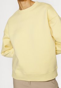 Jasnożółta bawełniana bluza z okrągłym dekoltem, opadającymi ramionami oraz ściągaczami przy rękawach i na dole. Minimalistyczny design, miękka tekstura.