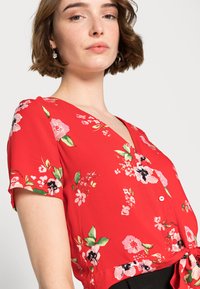 Blusa floral vermelha com mangas curtas, decote em V e bainha atada. Apresenta flores rosa e pretas com folhas verdes num tecido leve.