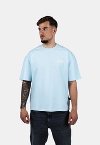 Světle modré bavlněné tričko s kulatým výstřihem, krátkými rukávy a malým bílým logem na levé hrudi. Volný střih, hladká textura.