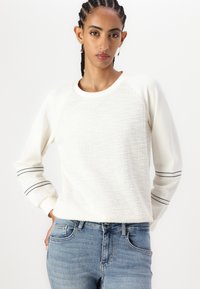 Sudadera blanca con un frente texturizado y mangas lisas con rayas negras, combinada con jeans azules. Diseño minimalista, ajuste casual.