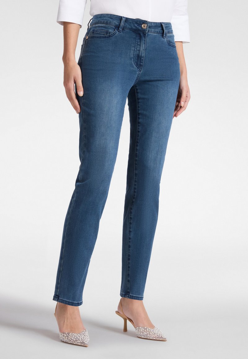 Mujer con jeans ajustados azules y camiseta blanca, de pie con tacones puntiagudos brillantes beige contra un fondo blanco.