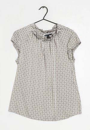 Blouse beige à manches courtes avec motif coquille blanc, col froufrou avec lien, suspendue sur un cintre noir sur fond blanc.