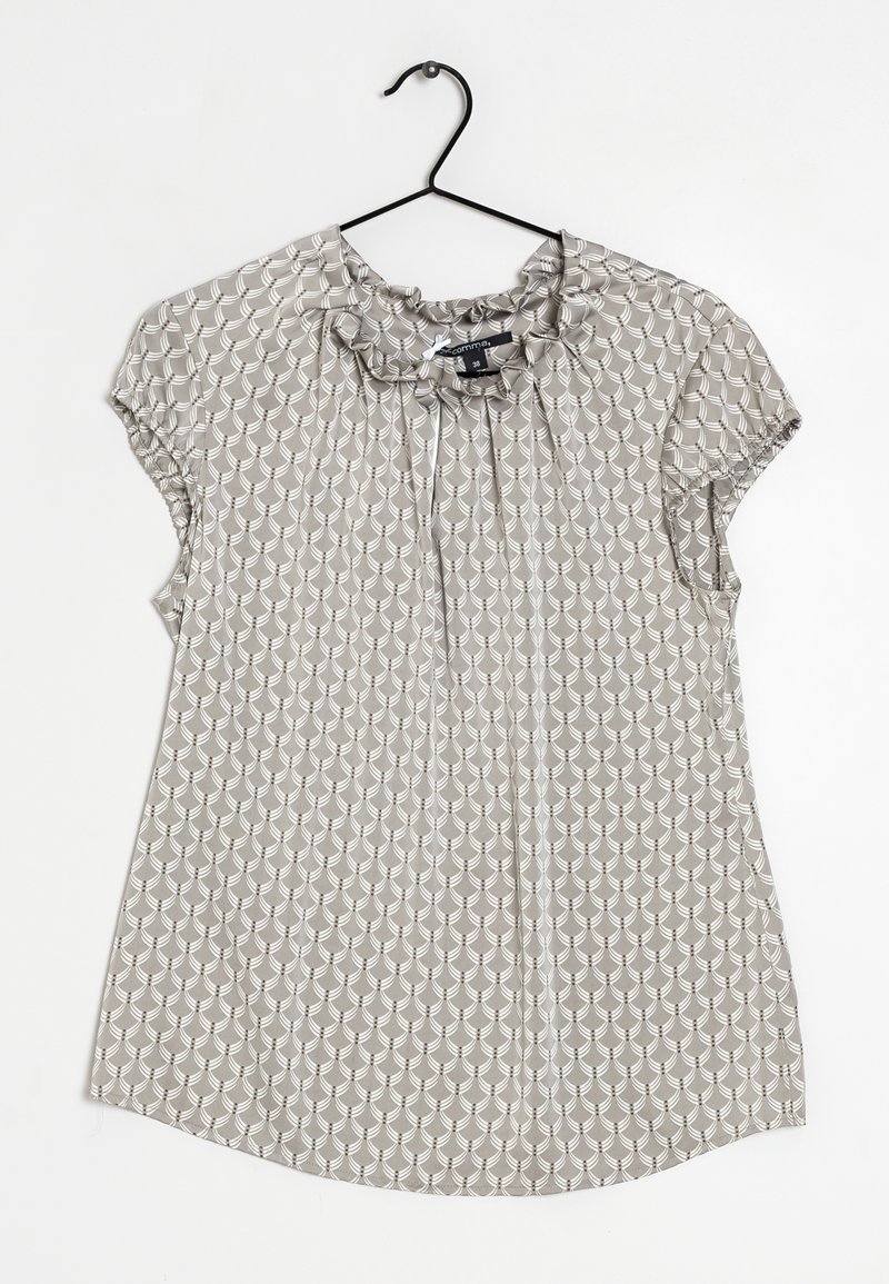 Blouse beige à manches courtes avec motif coquille blanc, col froufrou avec lien, suspendue sur un cintre noir sur fond blanc.