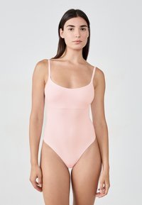 Kobieta z długimi ciemnymi włosami, ubrana w jasnoróżowy jednoczęściowy strój kąpielowy z cienkimi ramiączkami, stoi na tle jasnego, jednolitego tła.