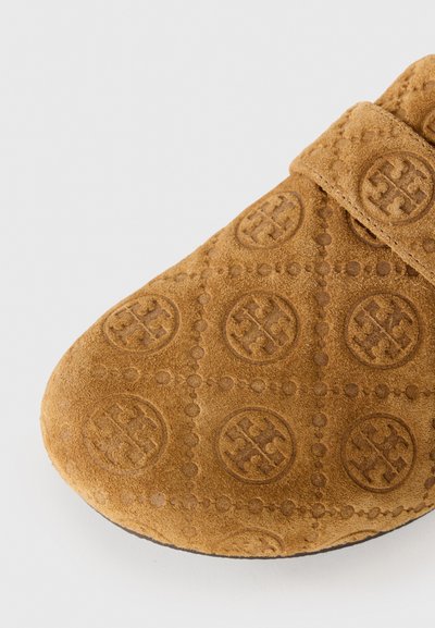 Toffel i beige mocka med präglat cirkulärt logomönster och stickad remdetalj på vit bakgrund.