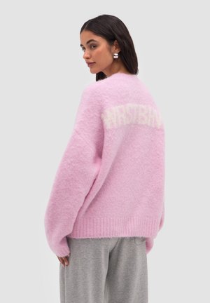Kvinde med mørkt hår iført sølvfarvede hoops øreringe og en lyserød, fluffy sweater med hvid tekst på ryggen, kombineret med grå bukser.
