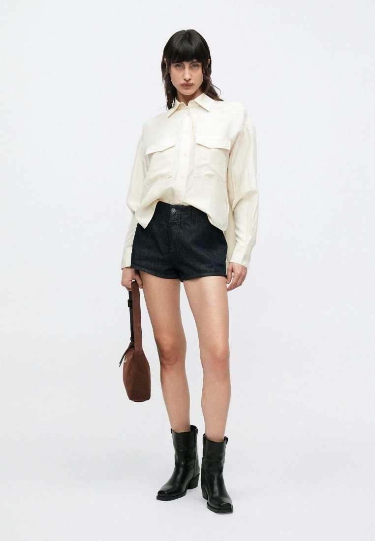 Camisa crema abotonada con dos bolsillos delanteros, combinada con pantalones cortos de mezclilla oscura y botines negros. Bolso marrón de hombro. Estilo sencillo y estructurado.